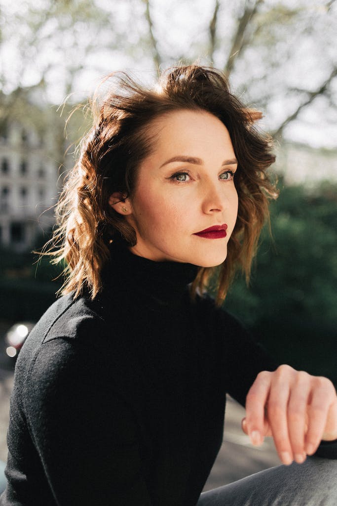Young contemplative female with makeup and wavy brown hair looking away while sitting in city in back lit